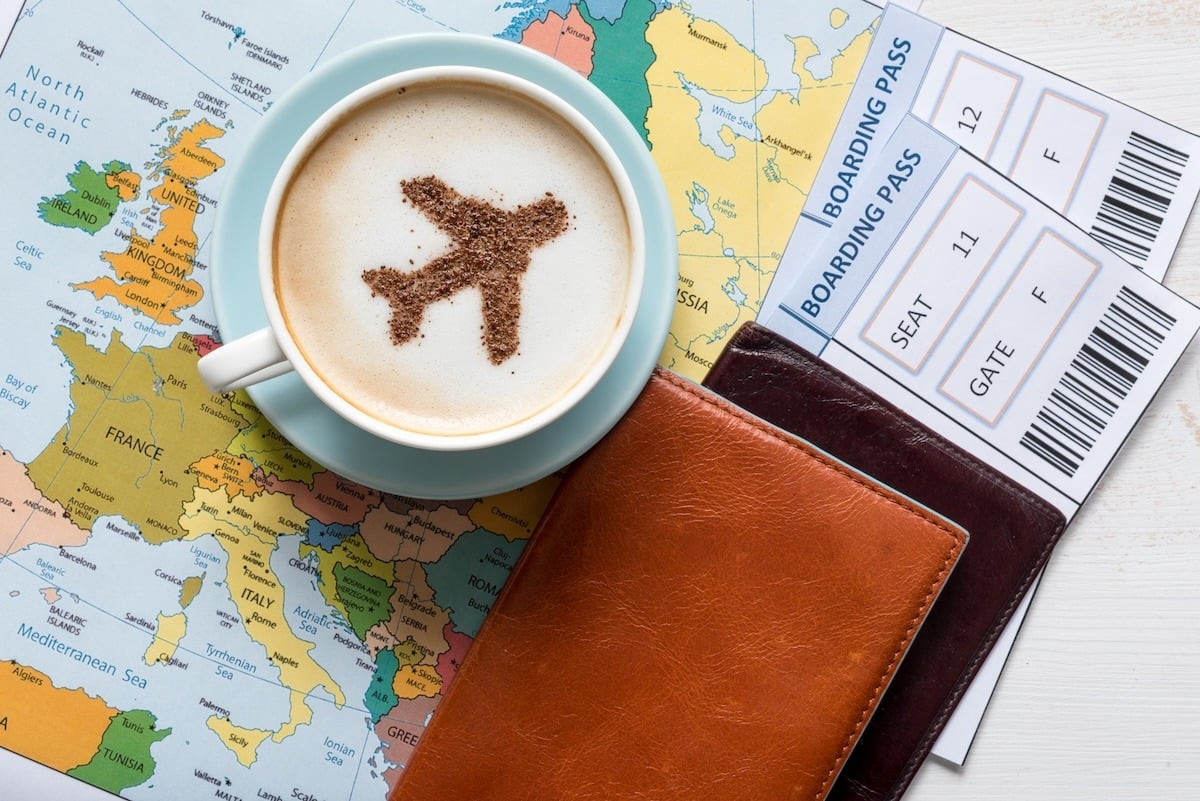 A cup of coffee with an airplane design on the foam sits on a world map, next to two boarding passes and a leather passport holder.