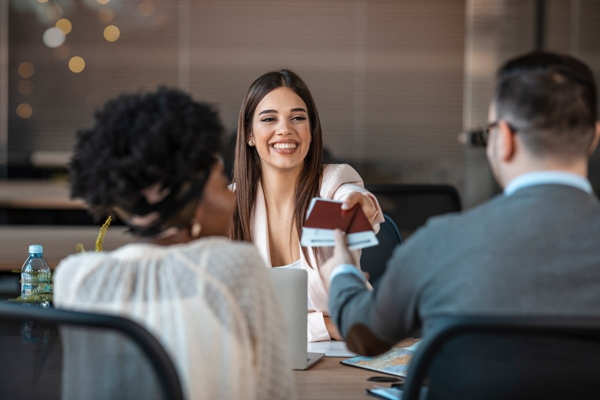 A smiling travel agent passes two tickets and passports across the table to her clients.