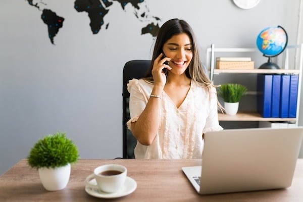 A smiling travel agent talks on the phone while working on her laptop, with a world map in the background.